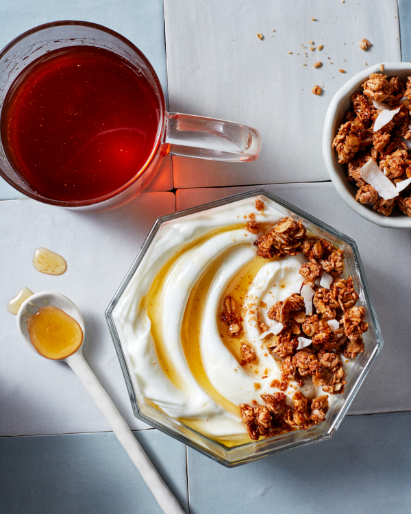 Un bol en verre octogonal de yaourt et de granola garni de sirop de gingembre sucré et épicé présenté avec une tasse en verre de sirop, un bol de granola et une cuillère à mélanger.