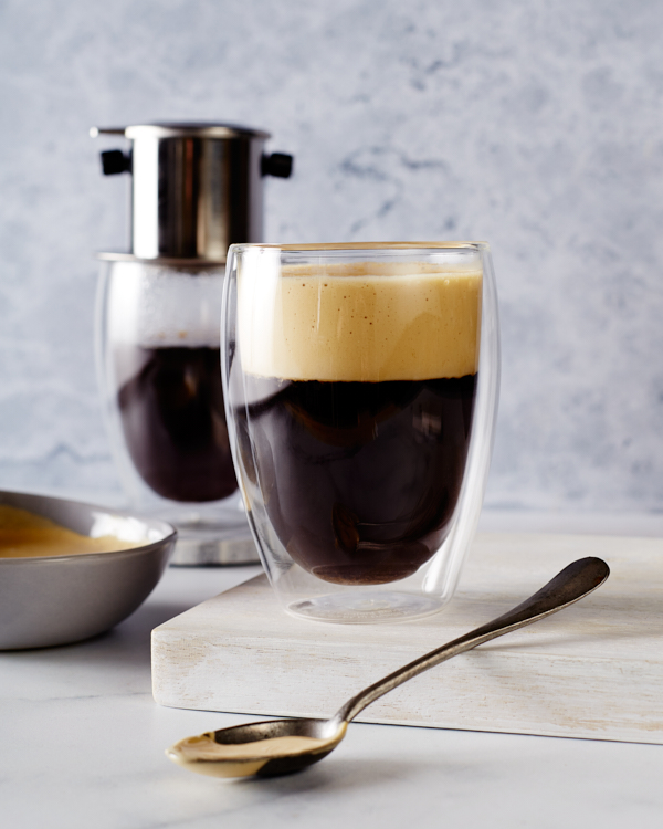 Un verre de café vietnamien à la mousse d'œuf montré avec un bol de mousse d'œuf, un verre de café et un équipement pour faire du café