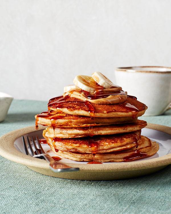 Une table dressée avec un plateau de crêpes au beurre d'arachide, du beurre, un bol de cacahuètes hachées, du beurre d'arachide, une tasse de café, des couverts, une carafe de sirop et deux assiettes avec des crêpes, du sirop et du beurre.