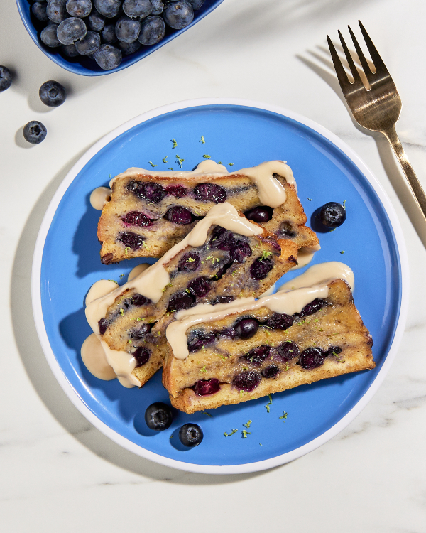 Trois tranches de pain doré cuites dans un moule avec des bleuets glacées d'érable sur une assiette bleue et blanche, présentées avec un plat de bleuets et une fourchette dorée.