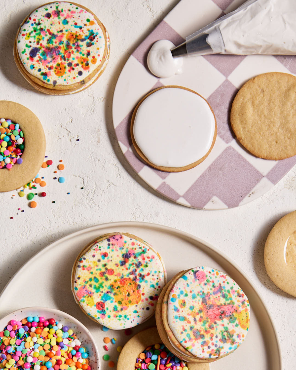  Trois biscuits en forme de piñata glacés, présentés avec deux biscuits incomplets remplis de confettis arc-en-ciel et de vermicelles, un bol de confettis arc-en-ciel et de vermicelles, ainsi qu'une poche à douille remplie de glaçage blanc qui fuit, le tout disposé sur un plateau à carreaux blancs et mauves.