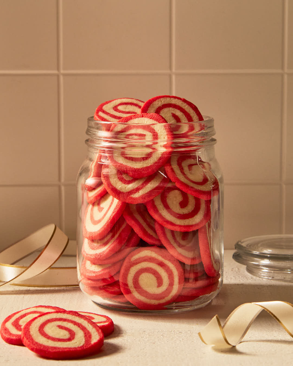 Un grand bocal en verre rempli de biscuits en spirale aux couleurs rouge et blanche sur un comptoir de cuisine devant un dosseret en carreaux de céramique de couleur crème, présenté avec le couvercle en verre, un ruban blanc et doré, et trois biscuits posés sur le comptoir.