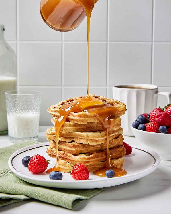 Du sirop versé de haut sur une pile de gaufres de babeurre dans une assiette blanche sur un comptoir de cuisine, présenté servi avec un bol de fraises et de bleuets, une tasse de lait et une tasse de café.