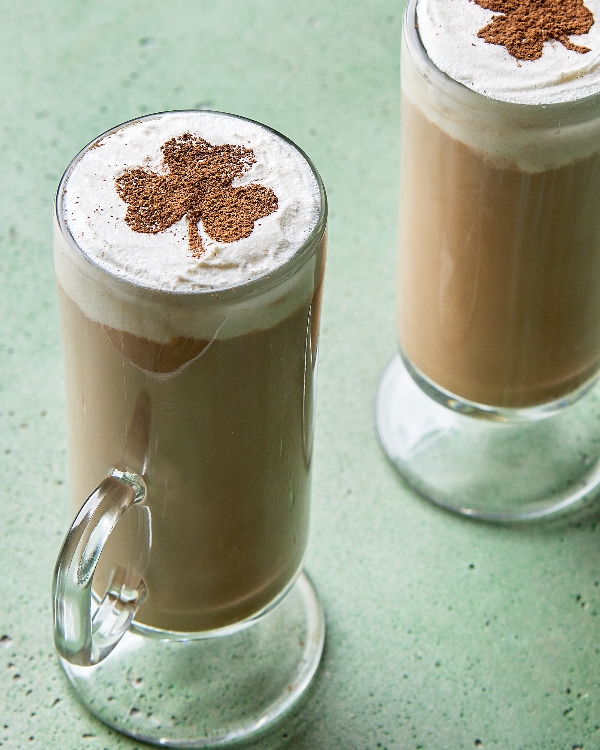 Deux lattes au whisky irlandais servis dans de grands verres transparents, surmontés de crème mousseuse et d'une pincée de cannelle en forme de trèfle, parfaits pour les célébrations de la Saint-Patrick, présentés devant un fond vert texturé.
