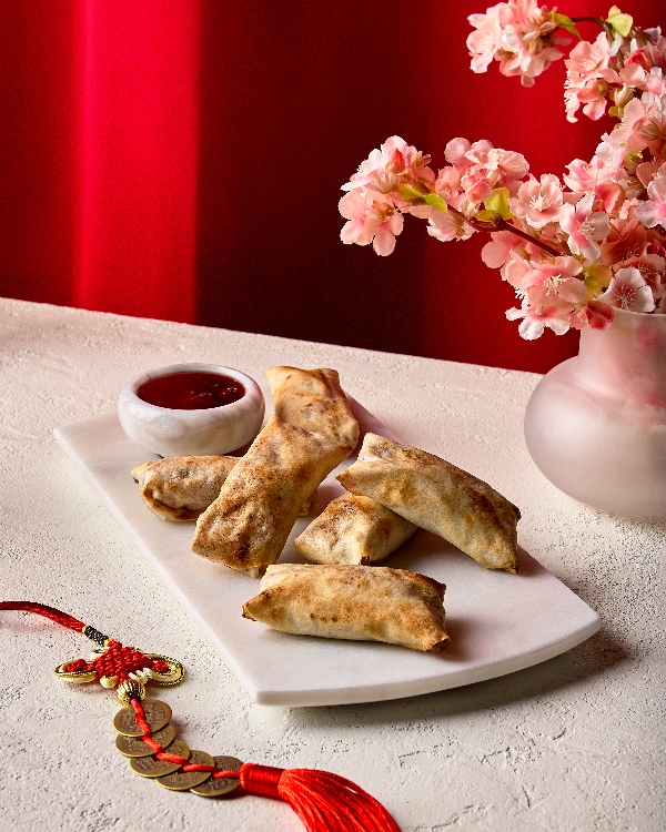 Rouleaux de printemps dorés aux pommes servis sur un plateau de marbre blanc avec un petit bol de sauce rouge. La mise en scène est rehaussée par des fleurs roses dans un vase et un nœud chinois rouge traditionnel avec des pièces d'or, le tout sur un fond rouge vif.