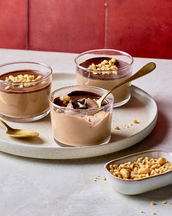  Trois poudings crémeux au chocolat et au beurre d’arachides dans des pots de verre, couverts de couches de chocolat craquant et d’arachides hachées. Une portion est partiellement mangée, révélant la texture soyeuse du pouding. Une cuillère dorée est déposée dans le pot. Les poudings sont disposés sur un plateau rond de couleur neutre avec un petit plat d’arachides hachés, le tout sur un fond rouge