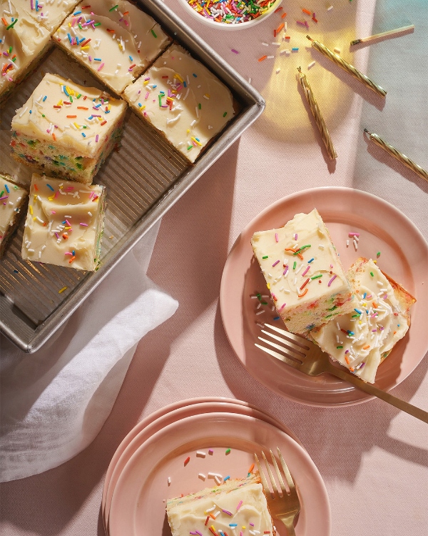 Un gâteau en plaque givré coupé en carrés décorés de vermicelles colorés, certains disposés dans un plat de cuisson et d'autres servis dans des assiettes roses avec des fourchettes dorées, ainsi que des vermicelles éparpillés et des bougies d'anniversaire sur une surface rose pâle.