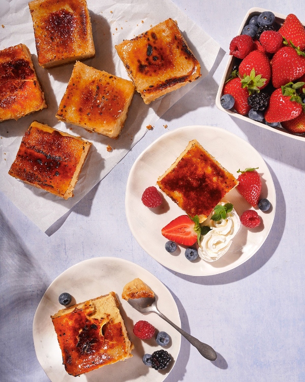 Une vue de dessus de portions individuelles de crème brûlée avec une croûte de sucre caramélisé, certaines nature sur du papier parchemin, et d'autres garnies de petits fruits frais et de crème fouettée sur de petites assiettes. Un bol de petits fruits mélangés se trouve dans le coin supérieur droit, et une cuillère repose sur un des desserts servis.