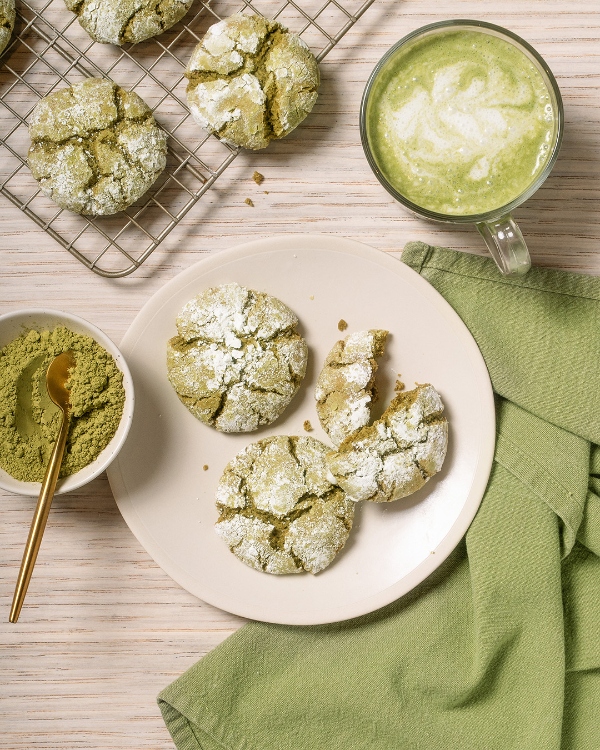 Une vue de dessus de biscuits craquelés au matcha sur une assiette, certains brisés, à côté d'un bol de poudre de matcha avec une cuillère dorée, un latté au matcha dans une tasse en verre, et d'autres biscuits sur une grille, le tout sur une surface en bois clair avec une serviette verte.