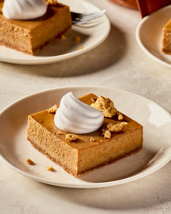 Une barre citrouille, bourbon et érable salé sur une assiette de couleur crème. La barre est garnie d’un tourbillon de crème fouettée et saupoudrée de morceaux de crumble doré. D’autres assiettes remplies de barres garnies apparaissent en arrière-plan, le tout sur une table décorée de tons neutres et baignée d’une lumière naturelle chaude.