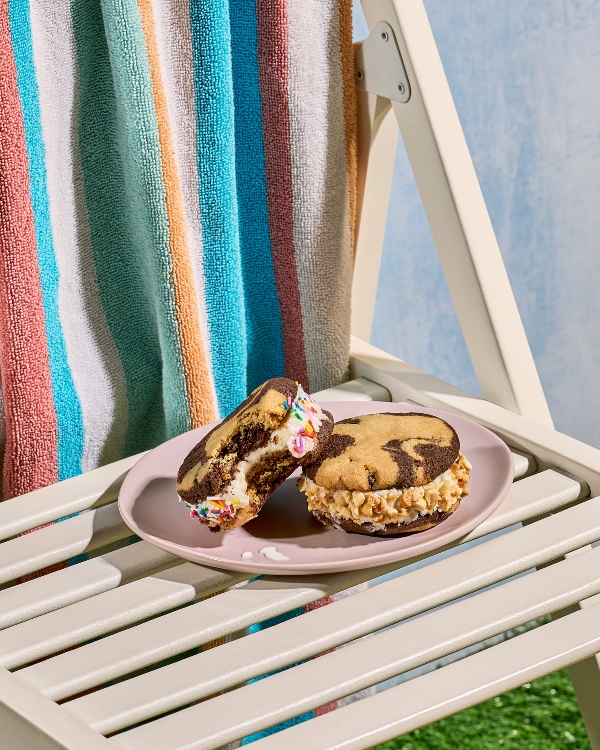 Deux sandwichs à la crème glacée marbrée au chocolat et à la vanille sont sur une assiette rose pâle, posée sur une chaise longue blanche à lattes. Un des sandwichs est roulé dans des vermicelles de couleur et a une bouchée manquante, l'autre est dans des noix hachées. Une serviette rayée colorée est accrochée au dossier de la chaise.