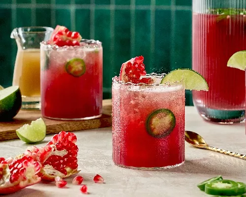 Trois verres de Fizz Épicé à la Grenade et au Gingembre, d'une couleur rouge vibrante avec des tranches de jalapeño frais. Les boissons sont garnies de quartiers de lime et de graines de grenade. Une carafe remplie de la même boisson se trouve en arrière-plan, aux côtés de quartiers de lime, de morceaux de grenade et d'une petite carafe de sirop de gingembre. La scène se déroule sur un fond de tuiles vert émeraude.