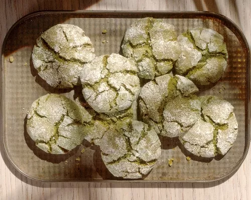 Une fournée de biscuits craquelés au matcha fraîchement sortis du four qui refroidissent sur une plaque à biscuits, montrant leurs surfaces craquelées et leur enrobage de sucre en poudre.