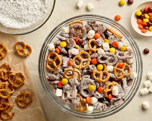 Vue du dessus d’un grand bol en verre rempli de Puppy Chow, une collation à base de céréales enrobées de chocolat et de bretzels saupoudrés de sucre à glacer. Autour du bol, il y a des bretzels éparpillés sur du papier parchemin, un récipient contenant du sucre à glacer et un petit plat rempli de pépites de chocolat. 