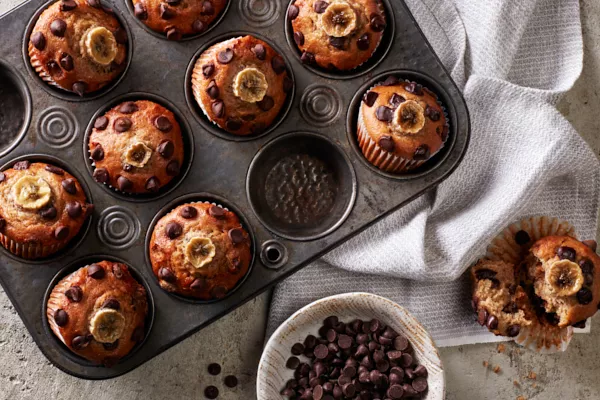 Muffins végétaliens à la banane et aux pépites de chocolat dans un moule à muffins, avec un muffin retiré et ouvert en deux, et un bol de pépites de chocolat.