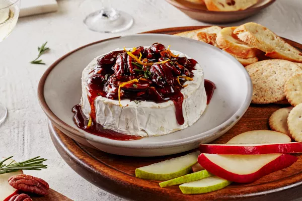  Brie cuit au four, garni de noix de pécan confites épicées et de sirop de Cassonade Foncée et décoré de zeste d'orange et de romarin, présenté sur une assiette sur un plateau de service avec des craquelins, des crostinis et des tranches de poires et de pommes.