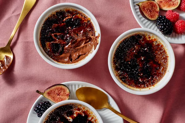  Trois ramequins de crème brûlée au chocolat noir salé, dont un partiellement mangé, présentés avec des cuillères dorées, des figues et des baies, sur une nappe de couleur rose. 
