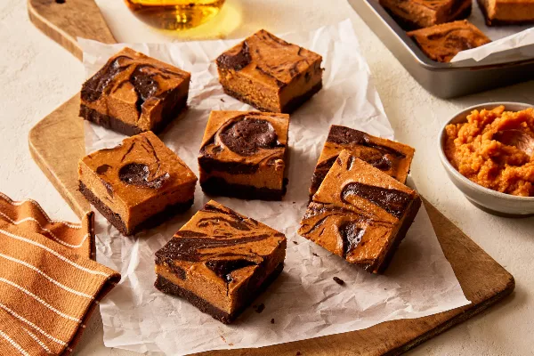 Sept brownies au cheesecake à la citrouille sur une planche à découper en bois doublée de papier parchemin, présentés avec un moule à brownies et un bol de purée de citrouille.
