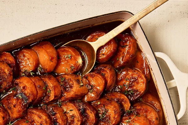 Des patates douces confites tranchées présentées dans un plat à gratin avec une cuillère de service, garnies d'herbes et montrant une caramélisation visible.