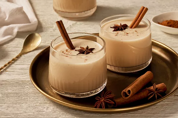 Deux verres tumblers de lait de poule sans œufs garnis d'anis, de cannelle, et de bâtons de cannelle, présentés sur un plateau doré avec de l'anis et des bâtons de cannelle, et une cuillère à mélanger dorée à côté.