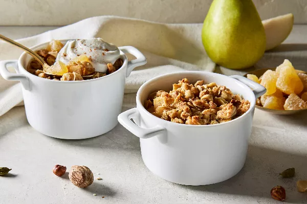 Deux bols blancs pour une portion de crumble poire-gingembre, l'un avec une touche de crème fouettée et une cuillère dorée, présentés avec une poire entière et un petit plat de morceaux de gingembre.