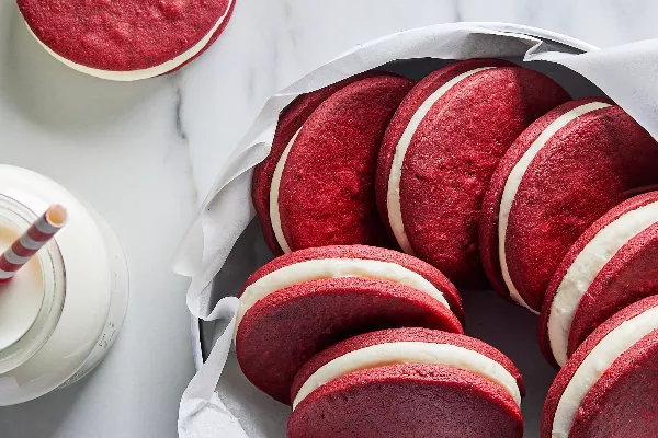 Des biscuits sandwich velours rouge présentés dans un bol tapissé de papier parchemin à côté d'un bocal en verre de lait avec une paille en papier.
