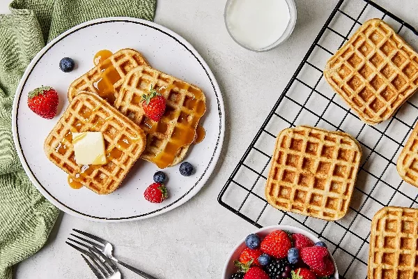 Trois gaufres de babeurre dans une assiette servies avec une noix de beurre et des fraises et des bleuets, présentées sur un comptoir de cuisine avec d'autres gaufres sur une grille de refroidissement, un verre de lait et un bol de baies mélangées.