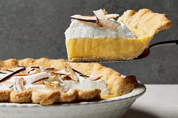 Un morceau de tarte à la crème de coco présentée de côté sur une pelle à tarte, fraîchement coupée d'une tarte entière dans un plat à tarte blanc décoratif, garnie de crème fouettée et de copeaux de noix de coco.