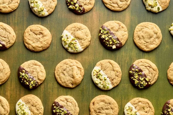 Biscuits sans pépites de chocolat disposés en quadrillage sur une surface en bois vert. Certains biscuits ne sont pas décorés, tandis que d’autres sont à moitié trempés dans du chocolat blanc ou noir et saupoudrés de pistaches hachées. 