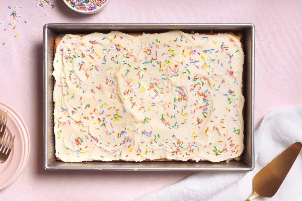 n gâteau rectangulaire givré recouvert de vermicelles colorés repose dans un plat de cuisson en métal sur une surface rose pâle, avec un petit bol de vermicelles supplémentaires, des assiettes et des fourchettes, et une pelle à gâteau dorée à proximité.
