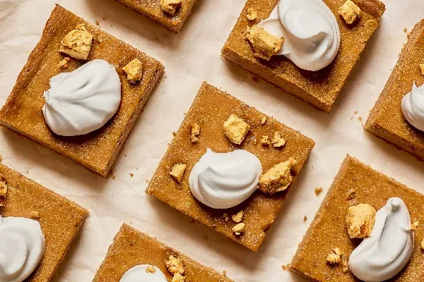 Vue aérienne de barres à la citrouille méticuleusement disposées sur du papier parchemin. Chaque barre est couronnée d’un tourbillon de crème fouettée et saupoudrée de quelques morceaux de crumble doré et croustillant. Les barres sont d’une riche teinte orange foncé.