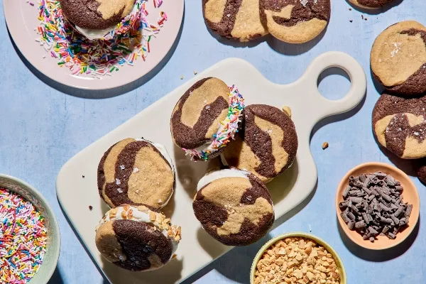Un plateau de sandwichs à la crème glacée marbrée au chocolat et à la vanille est posé sur une planche de service blanche. Certains sont roulés dans des vermicelles de couleur ou des noix hachées. D'autres biscuits et des bols de garnitures entourent la scène sur une surface bleu pâle.
