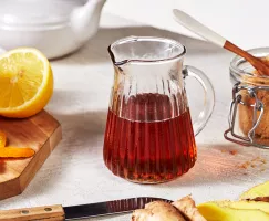 Un bol en verre octogonal de yaourt et de granola garni de sirop de gingembre sucré et épicé présenté avec une tasse en verre de sirop, un bol de granola et une cuillère à mélanger.