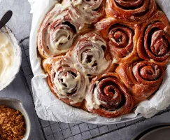 Un plat de cuisson rond tapissé de papier sulfurisé et rempli de petits pains à la cannelle, dont la moitié est glacée, présenté avec un bol de glaçage et un bol de Cassonade Foncée.