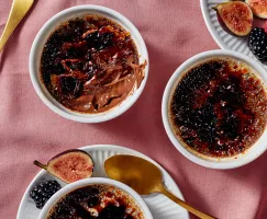  Trois ramequins de crème brûlée au chocolat noir salé, dont un partiellement mangé, présentés avec des cuillères dorées, des figues et des baies, sur une nappe de couleur rose. 