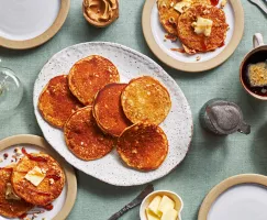 Une table dressée avec un plateau de crêpes au beurre d'arachide, du beurre, un bol de cacahuètes hachées, du beurre d'arachide, une tasse de café, des couverts, une carafe de sirop et deux assiettes avec des crêpes, du sirop et du beurre.
