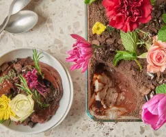 Pouding au chocolat saupoudré de miettes de biscuits et garni de fleurs comestibles dans un plat à four en verre, présenté avec une portion de pudding dans un bol et des cuillères.