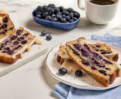 Deux tranches de pain doré cuites dans un moule avec des bleuets glacées d'érable, présentées avec une tasse de café, une assiette de bleuets et un pain partiellement tranché
