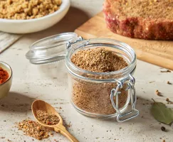 Un bocal en verre avec un couvercle rempli d'épices sucrées pour steak à la montréalaise est présenté avec un steak cru assaisonné sur une planche à découper, un bol de sucre brun et un bol d'épices. 