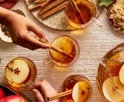 Quatre tasses en verre de cidre de pomme chaud garnies de tranches de pomme, de clous de girofle et de bâtonnets de cannelle, présentées sur une table avec une carafe de cidre, un bol de pommes et un bol de sucre jaune doré, et des mains tendues vers les tasses.