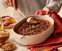 Une femme portant un chandail sert une casserole de patates douces garnie d'une branche de romarin depuis un plat de cuisson, présentée avec une salade de radicchio et d'agrumes, des verres de vin et des bols de sucre jaune doré et de sucre Demerara.