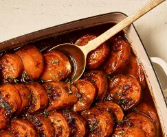 Des patates douces confites tranchées présentées dans un plat à gratin avec une cuillère de service, garnies d'herbes et montrant une caramélisation visible.