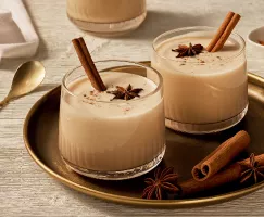 Deux verres tumblers de lait de poule sans œufs garnis d'anis, de cannelle, et de bâtons de cannelle, présentés sur un plateau doré avec de l'anis et des bâtons de cannelle, et une cuillère à mélanger dorée à côté.