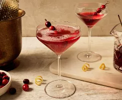 Un verre à pied rempli d'un cocktail pétillant à la canneberge, garni d'une brochette de canneberges et de zeste de citron, présenté avec une carafe de cocktail à la canneberge, un verre à pied de cocktail à la canneberge non pétillant, du zeste de citron, et une bouteille de vin mousseux dans un seau à glace en laiton.