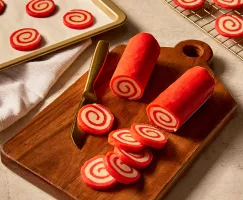 Deux rouleaux de pâte à biscuits en spirale aux couleurs rouge et blanche, posés sur une planche à découper en bois avec plusieurs tranches coupées, et d'autres tranches sur une plaque de cuisson, ainsi que des biscuits cuits refroidissant sur une grille en métal