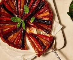 Une tarte tatin aux carottes et aux panais reposant sur du papier sulfurisé sur une planche à découper, présentée avec une tranche partiellement retirée à l'aide d'une pelle à tarte, garnie de feuilles de menthe fraîche.