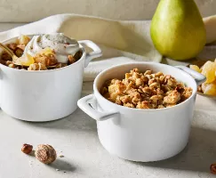 Deux bols blancs pour une portion de crumble poire-gingembre, l'un avec une touche de crème fouettée et une cuillère dorée, présentés avec une poire entière et un petit plat de morceaux de gingembre.