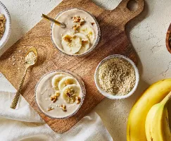  Vue de dessus de deux grands verres de smoothie à la banane, avoine et tahini, présentés sur une planche à découper en bois avec des bols de sucre jaune doré, de graines de sésame et d'avoine sèche, accompagnés de deux bananes non pelées.