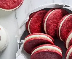 Des biscuits sandwich velours rouge présentés dans un bol tapissé de papier parchemin à côté d'un bocal en verre de lait avec une paille en papier.