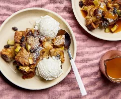 Une portion de pudding de pain aux pommes dans une assiette avec des raisins secs et des morceaux de pomme visibles, servie avec de la glace à la vanille, présentée avec une deuxième assiette de pudding de pain avec sauce au caramel et un pichet en verre de sauce.
